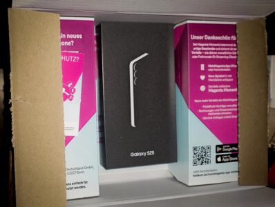 Offener Versandkarton, darin ein schwarzer Samsung Galaxy S25 Karton zwischen Magenta Telekom Unterlagen.
