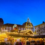 Weihnachtsmarkt in Darmstadt am Abend, beleuchtete Buden, Lichterketten und dekorierte Dächer, Menschen im Gedränge, blauer Himmel in der Dämmerung.