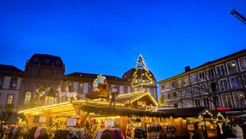 Weihnachtsmarkt in Darmstadt am Abend, beleuchtete Buden, Lichterketten und dekorierte Dächer, Menschen im Gedränge, blauer Himmel in der Dämmerung.