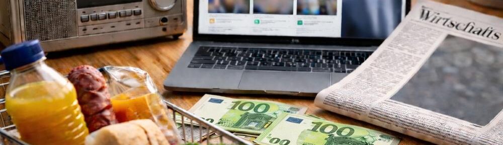 Radio, Laptop, Zeitung, Einkaufskorb und Euroscheine als Sinnbild für Konsumdruck und Dauerbeschallung, fotorealistisch per KI erstellt.