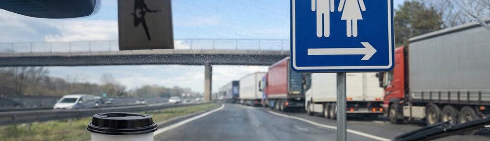 Blick aus dem Auto auf die Autobahn mit Kaffeebecher, Wischerblick, wartenden Lkw und WC-Schild am Rastplatz, auf Grundlage eines eigenen Fotos mit KI ergänzt.