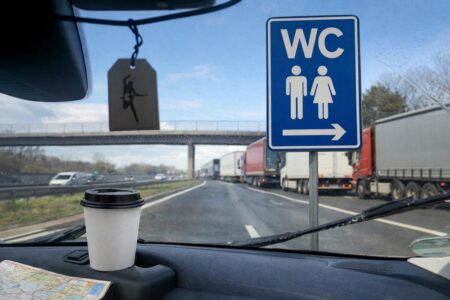 Blick aus dem Auto auf die Autobahn mit Kaffeebecher, Wischerblick, wartenden Lkw und WC-Schild am Rastplatz, auf Grundlage eines eigenen Fotos mit KI ergänzt.