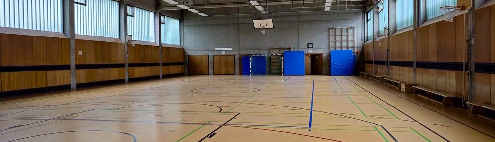 Leere Schulturnhalle mit Spielfeldlinien, Basketballkörben und blauen Matten.